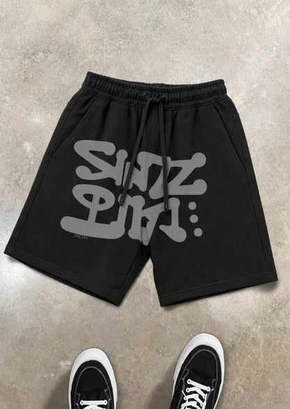 Black shorts with gray text on a concrete floor, paired with black sneakers.